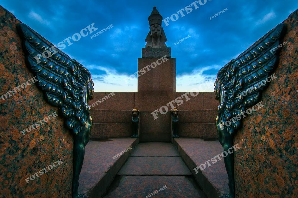 На фотографии изображены крылатые гранитные сфинксы, установленные на набережной Невы в Санкт-Петербурге. Их массивные каменные крылья и древний стиль создают ощущение величия и таинственности. Эти скульптуры, символизирующие связь Петербурга с древними цивилизациями, особенно эффектно выглядят на фоне сумеречного неба. Сфинксы являются одним из знаковых элементов городской архитектуры и привлекают как туристов, так и местных жителей.